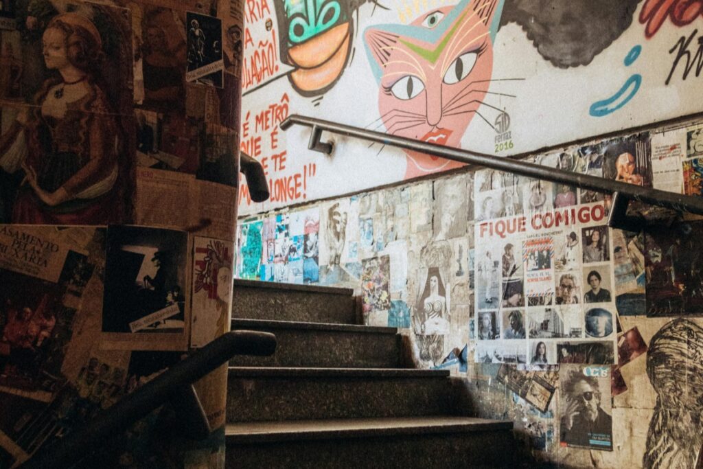 Esta imagem mostra uma escada cercada por paredes cobertas de cartazes, pôsteres e uma arte mural com um gato colorido. Há textos em português dispersos entre as imagens, como "FIQUE COMIGO". Quando se trata de "Como Impulsionar Seu Negócio com Anúncios no Facebook Brasil", se o seu negócio está ligado à arte, cultura ou evento local, imagens como esta podem ser muito eficazes. No Facebook Ads, você pode destacar a vibração e a criatividade de locais assim. Use hashtags como #ArteMuralBrasil, #CulturaLocal e #EventosCulturaisBrasil para alcançar o público-alvo correto. Isso pode ajudar a chamar a atenção dos usuários interessados em arte e cultura, aumentando a visibilidade do seu negócio e potencialmente levando a mais clientes e vendas.